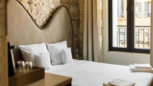 A hotel room with a stone wall, a bed with white linens, a wooden nightstand, and a window with a view of buildings. - Notre Dame City Residence (Nice)