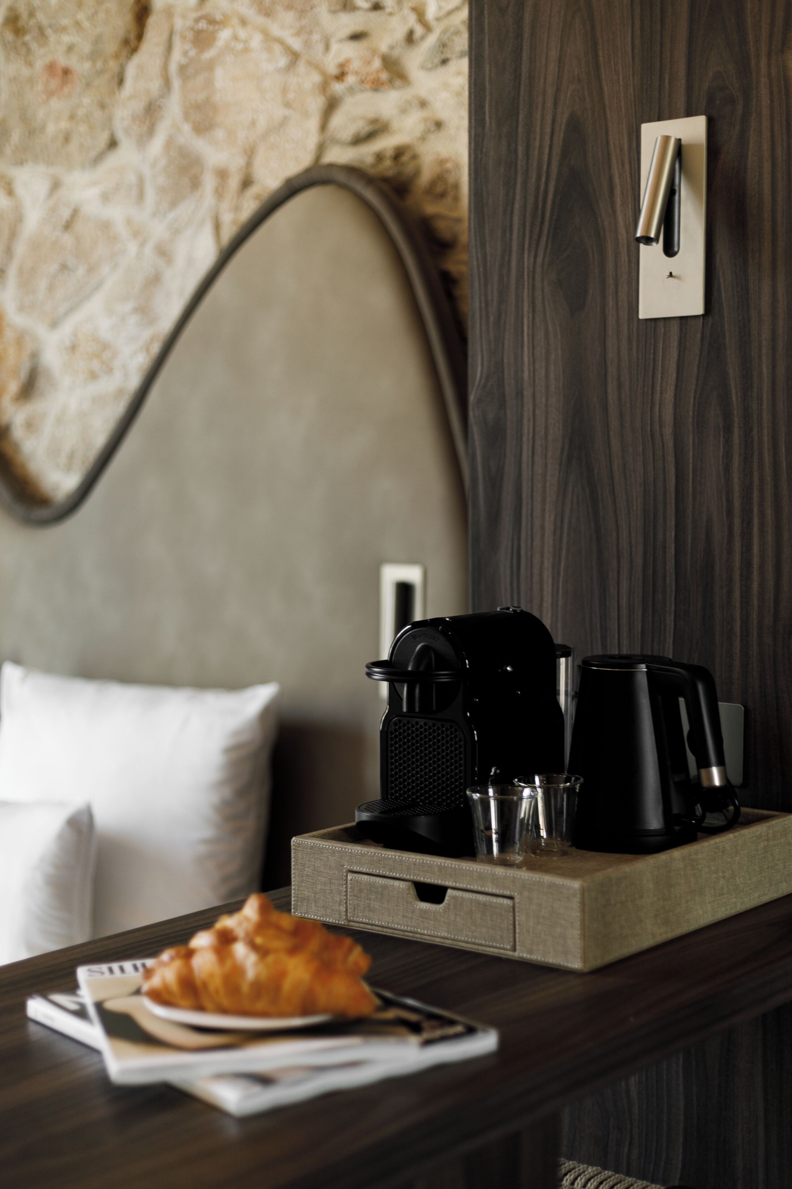 Hotelkamer met een stenen muur, een bed met hoofdeinde, een dienblad met koffiezetapparaat, een croissant en een krant.