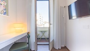 Une pièce comprenant un bureau, une chaise, une lampe et donnant sur un balcon avec une table et des chaises, accessible par une porte ouverte.