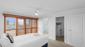 Ein Schlafzimmer mit Bett, Deckenventilator, Fenster mit Jalousien und begehbarem Kleiderschrank.