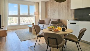 Un salon moderne comprenant un canapé, une table à manger avec des chaises, une machine à café et une vue sur la ville grâce à de grandes fenêtres.