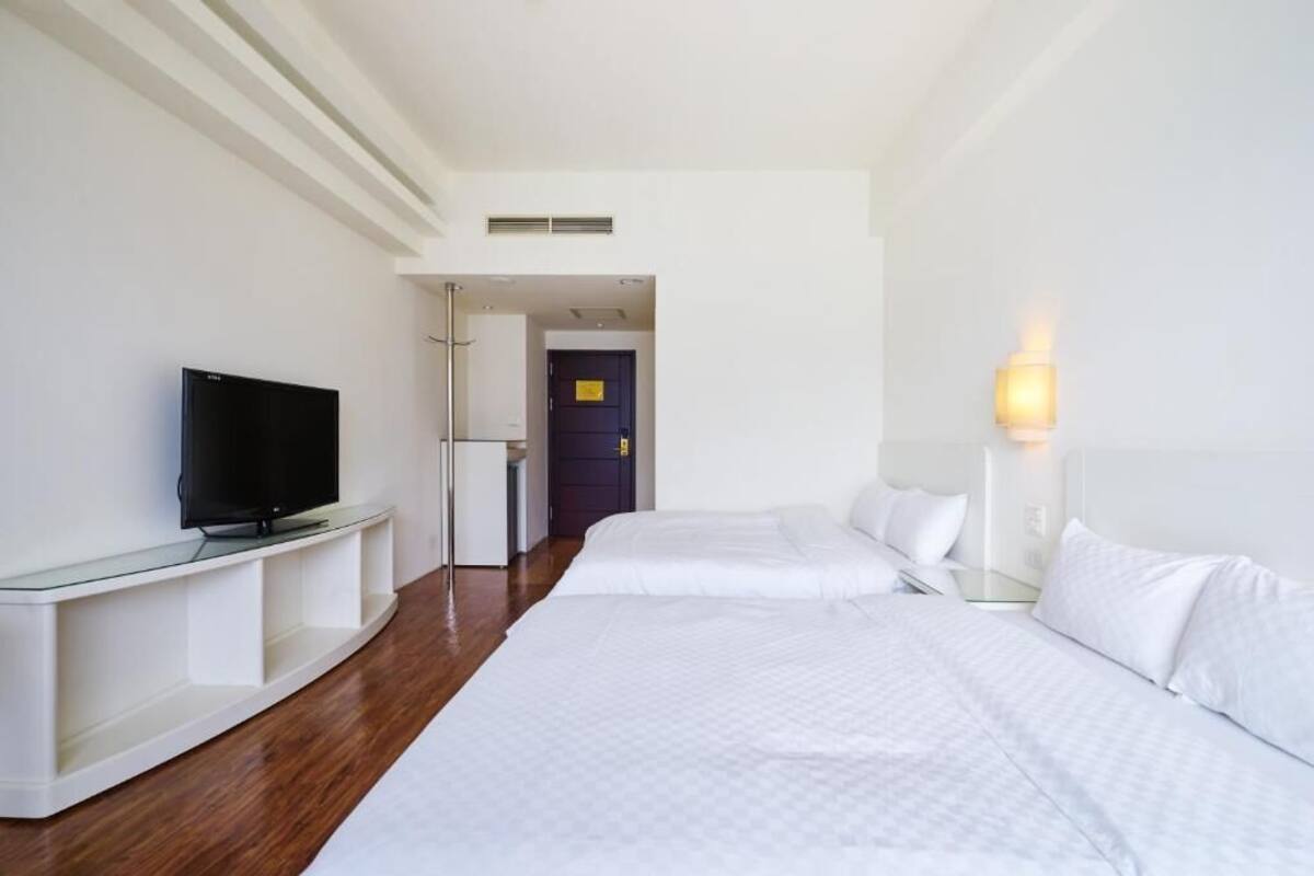A hotel room with a TV on a glass stand, two beds with white bedding, a bedside table with a lamp, and a door leading to another room.