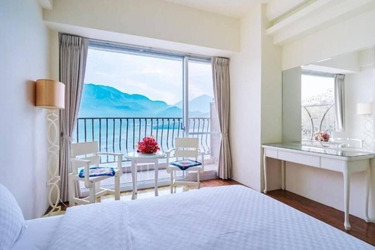 A hotel room with a bed, a desk, a chair, a small table with a vase, and a balcony with a view of mountains.