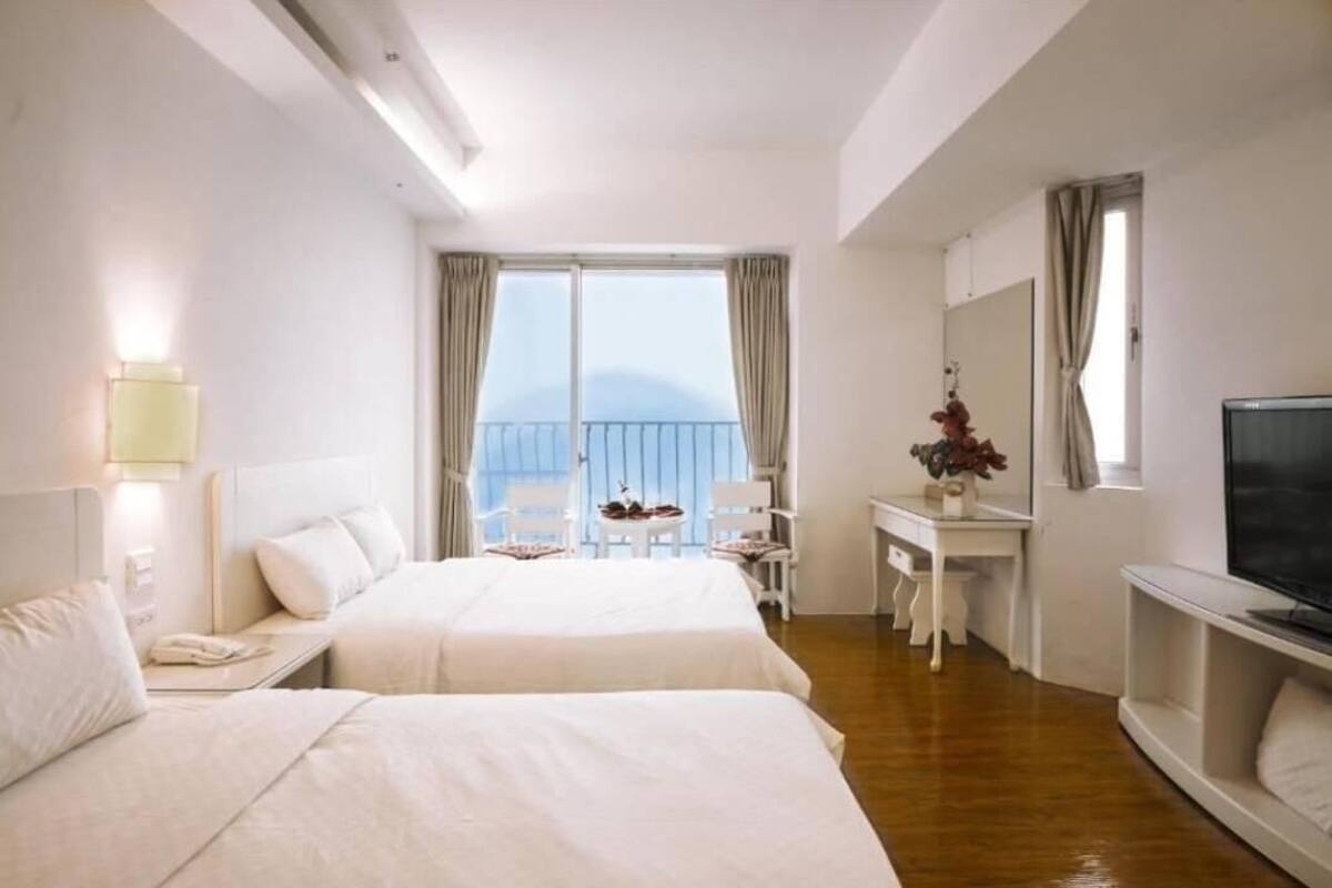 A hotel room with two beds, a desk, a chair, a TV, and a balcony with a view.