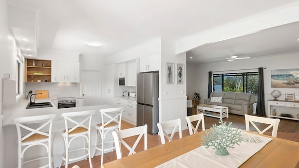 A modern kitchen with white cabinets, a dining area with white chairs, and a living room with a sofa and a large window. - Robins Place - Pet Friendly River Retreat (Empire Vale)