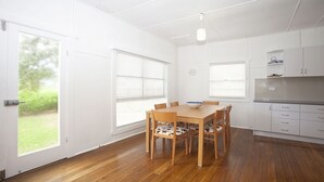 A modern kitchen with white cabinets, a wooden dining table, and a view of the outdoors through large windows. - My Blue Heaven Dog Friendly (Lennox Head)