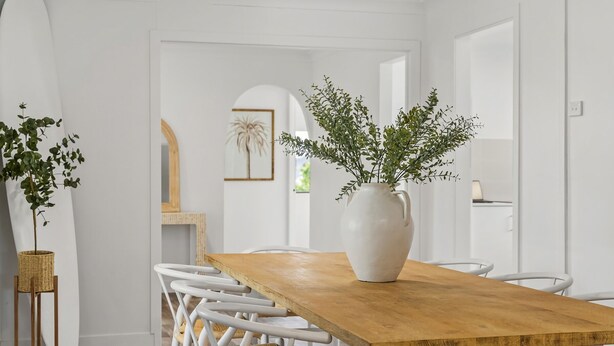 Un comedor con una mesa de madera, sillas blancas y un jarrón blanco con follaje.