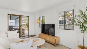 Una sala moderna con sofá, televisor y un comedor visible a través de una puerta de vidrio.