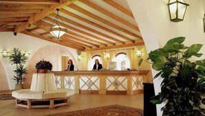 Eine Lobby mit einer Rezeption, zwei Mitarbeiterinnen an der Rezeption, einer großen Kübelpflanze, einem gemusterten Teppich und Holzbalken an der Decke.