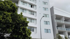 A multi-story building with balconies, a parking area, and a tree in the foreground.
