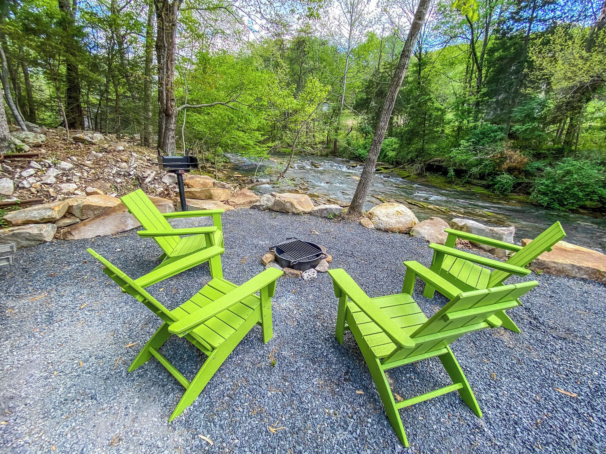 Un camping con cuatro sillas Adirondack verdes dispuestas alrededor de un brasero central, rodeado por una zona de grava con un arroyo y un bosque al fondo.