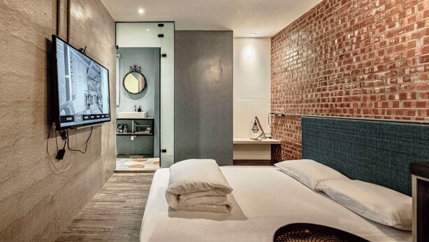 Quarto de hotel moderno com parede de tijolos, televisão de tela plana, cama com lençóis brancos, mesa de cabeceira e espelho circular.