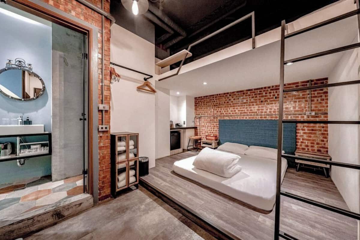 Una habitación de hotel moderna con pared de ladrillo decorativa, cama con ropa blanca, lámpara colgante y repisa de madera con toallas.