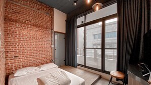 Um quarto com parede de tijolos, cama com roupa branca, janela com cortinas e um banquinho.