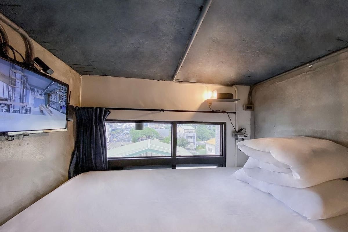 Una habitación con cama, televisor montado en la pared, ventana con vista a edificios y lámpara sobre la ventana.