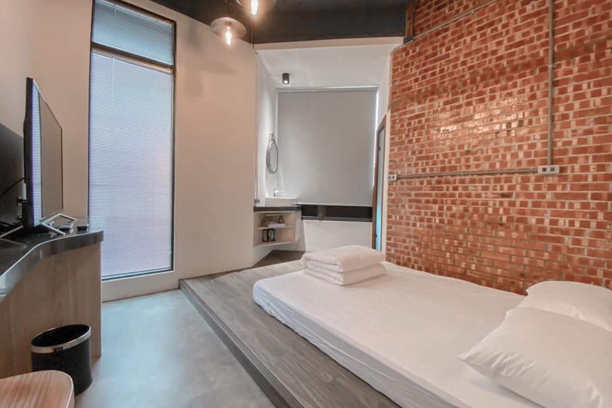 Una habitación de hotel moderna con una pared de acento de ladrillo, una cama grande, un televisor integrado y un área de ducha.