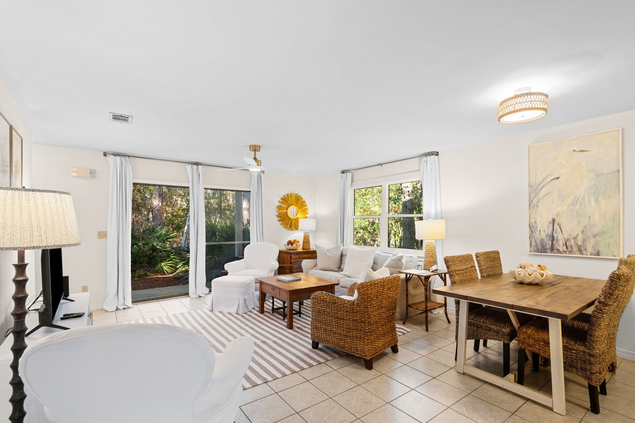 A spacious room with a large window, a dining table, wicker chairs, a sofa, and a TV.