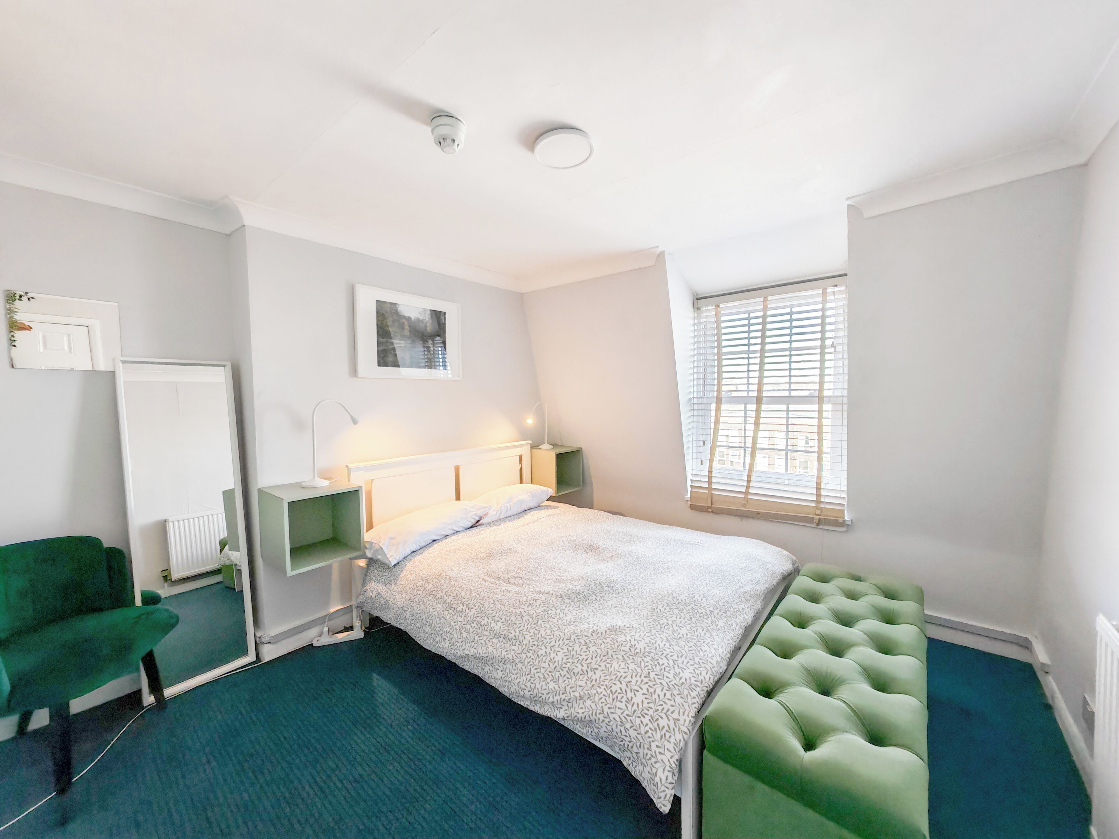 A bedroom with a bed, a green chair, a radiator, a window with blinds, and a ceiling light.