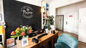 Una mesa de recepción con un letrero en la pared negro, una computadora portátil, un teléfono inteligente, un cartel con un código QR, un florero con flores, una silla azul y una estantería con libros y una planta.