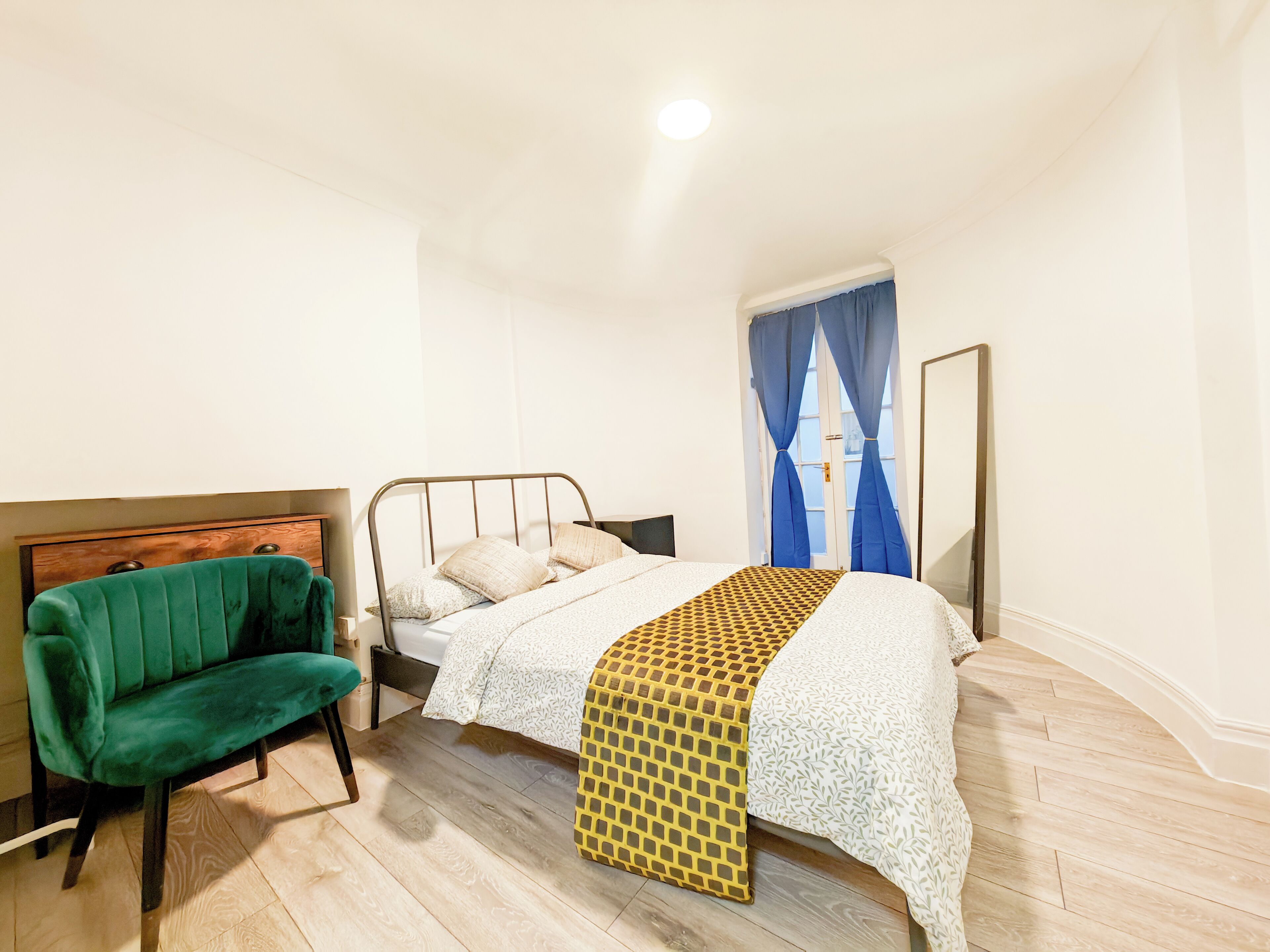 A bedroom with a bed, a green chair, a wooden headboard, a window with blue curtains, and a mirror.