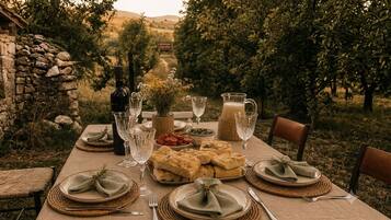 An outdoor dining table set with plates, glasses, and a bottle, surrounded by a stone wall and trees.