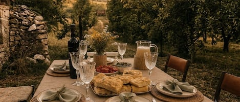 Tavolo da pranzo all'aperto apparecchiato con piatti, bicchieri e una bottiglia, circondato da un muro in pietra e alberi.