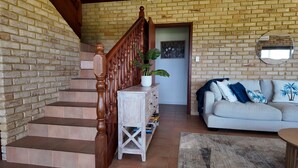 Una escalera con barandillas de madera, una consola blanca con una planta, un sofá gris con cojines decorativos y un espejo redondo en la pared.