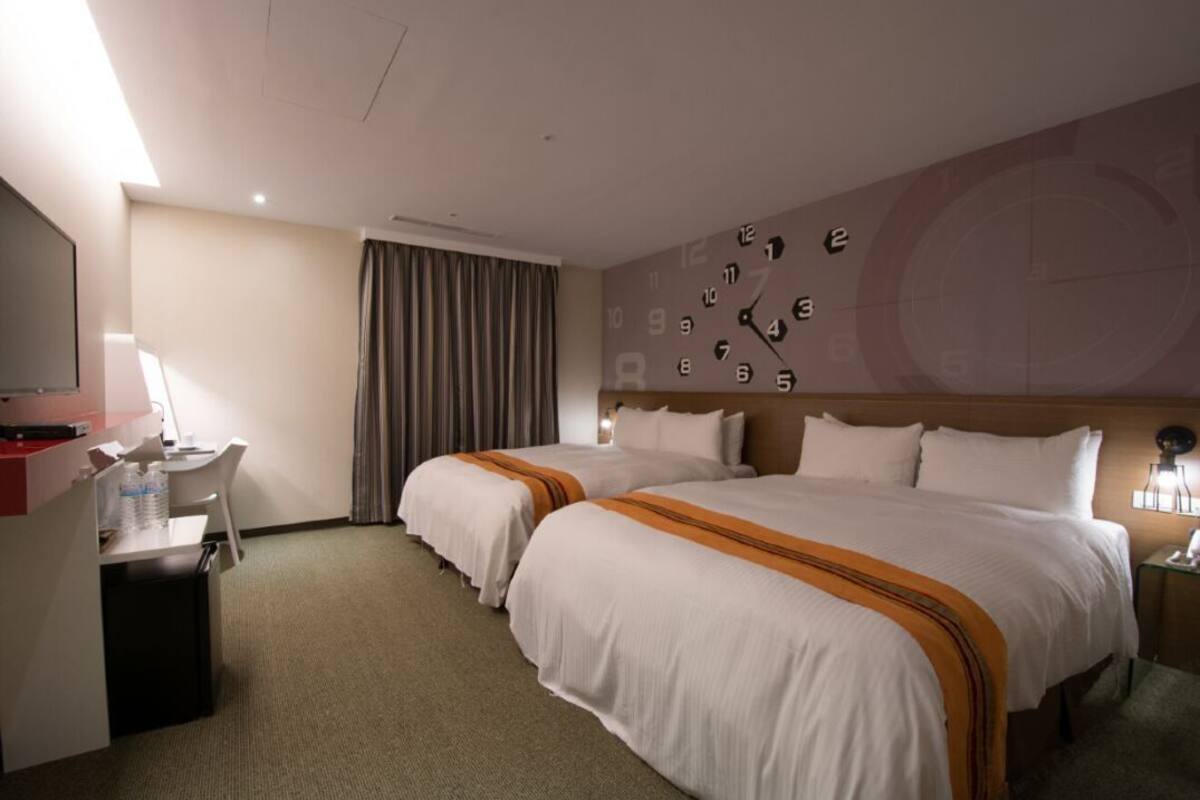 Ein Hotelzimmer mit zwei Betten, einem Schreibtisch, einem Fernseher und einer Wanduhr.