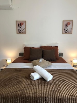 Ein ordentlich bezogenes Bett mit einer braun gestreiften Tagesdecke, zwei Kissen und einem gefalteten weißen Handtuch. Zwei Nachttischlampen und zwei Wandbilder sind vorhanden.