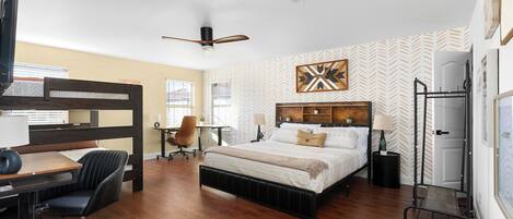 A bedroom with a bed, desk, chair, ceiling fan, and a wall with patterned wallpaper.
