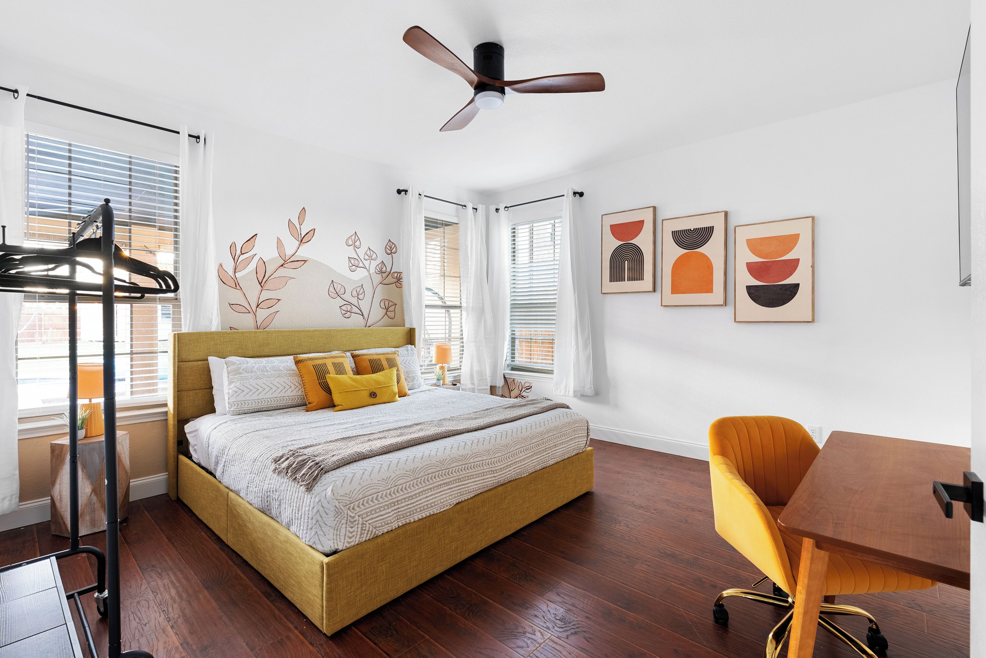 A bedroom with a bed, a desk, a chair, a ceiling fan, and artwork on the wall.