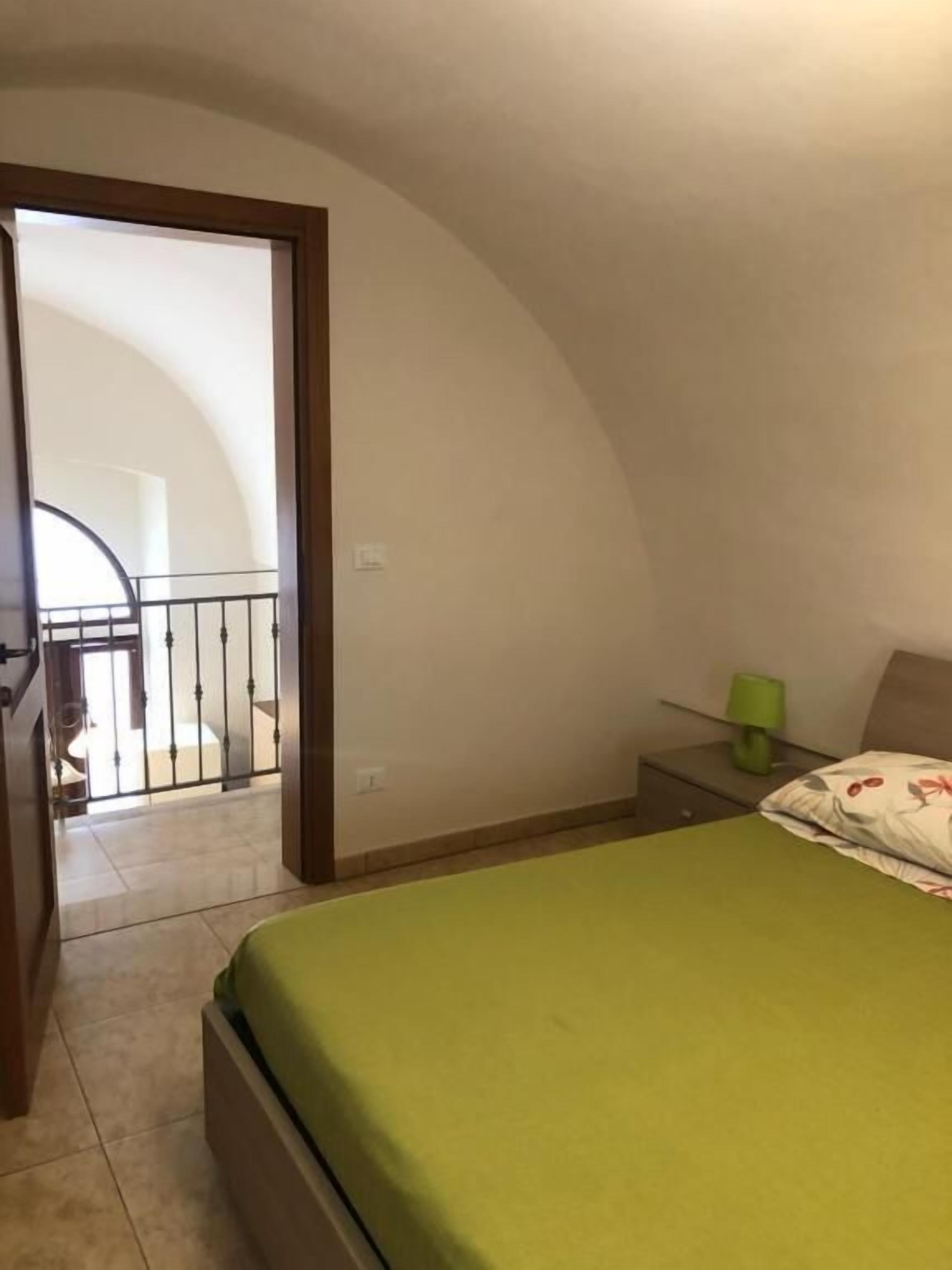 Quarto com cama, mesa de cabeceira, abajur verde e vista para uma varanda.