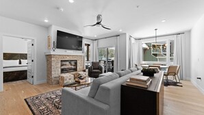 Un salon moderne doté d’une cheminée, d’un grand téléviseur à écran plat, d’un ventilateur de plafond et d’un coin repas offrant une vue sur l’extérieur.