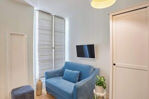 A room with a blue sofa, a small ottoman, a potted plant, a television on the wall, and a sliding door. - Charming Suite with AC - Center of Paris (Paris)