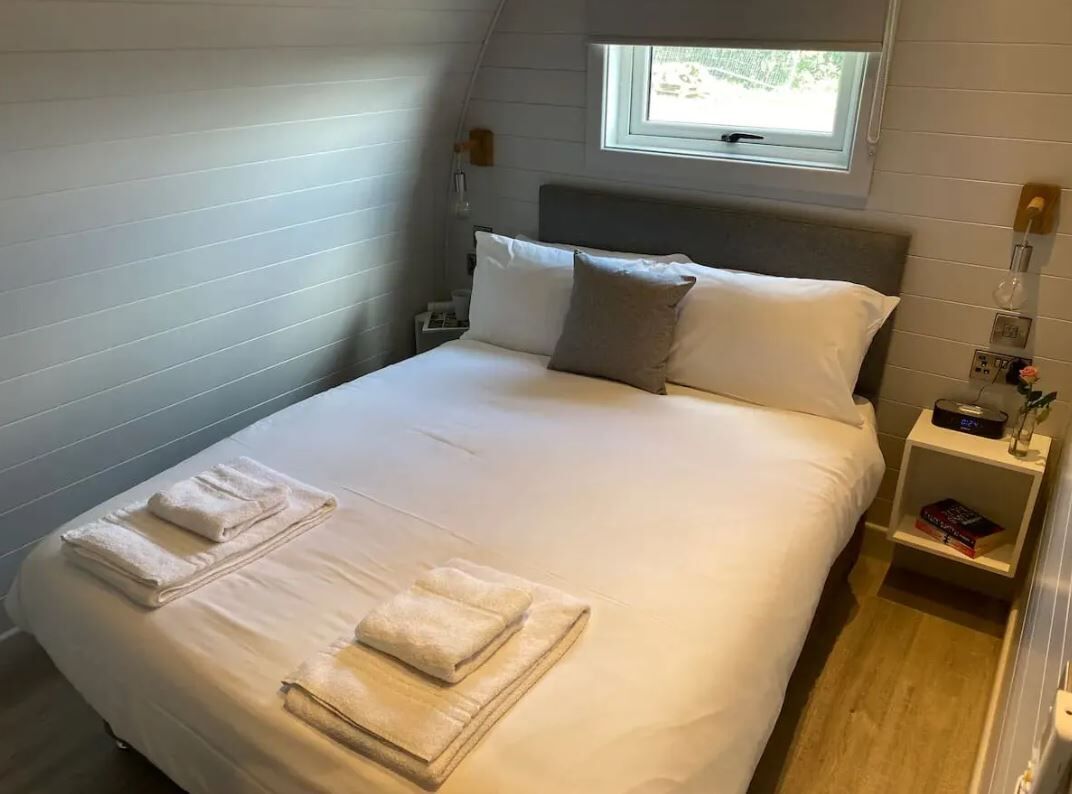 A neatly made bed with folded towels, a bedside table with a book, a nightstand with a lamp, and a window above the bed.