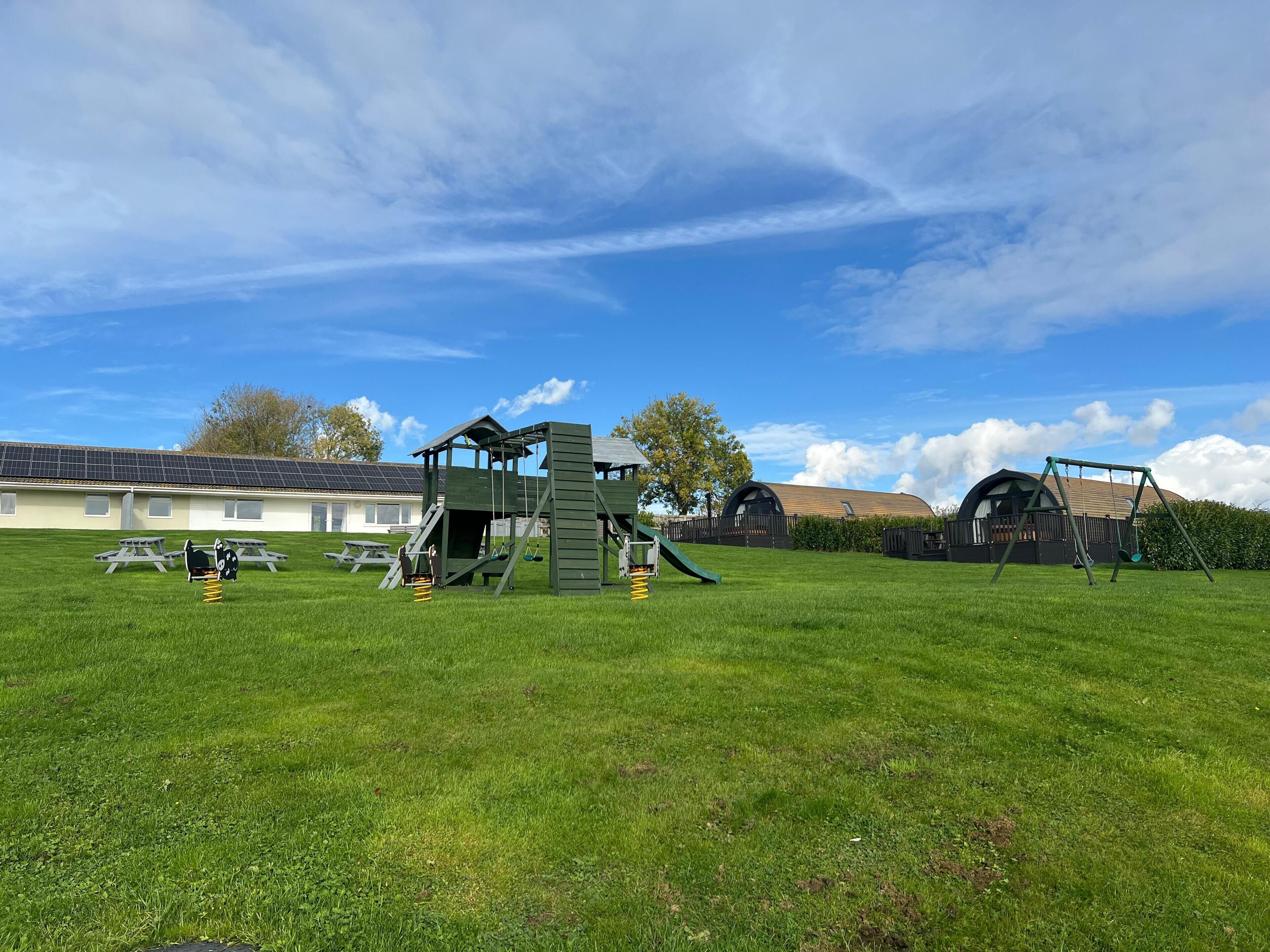 A playground with climbing structures and swings, a picnic table, and a building with a sloped roof in the background.