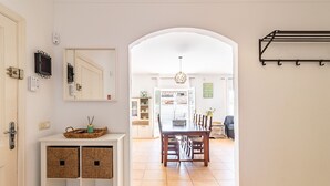 Um corredor leva a uma área de jantar com mesa e cadeiras de madeira, um aparador branco com cestos e um suporte fixado na parede.