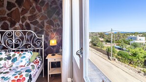 Uma varanda com parede de pedra, uma cama com colcha florida, uma mesa de cabeceira com abajur e vista para uma rua e edifícios.