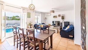Uma sala de estar ampla com uma mesa de jantar, cadeiras, um sofá e uma TV. Uma piscina é visível pela porta aberta.