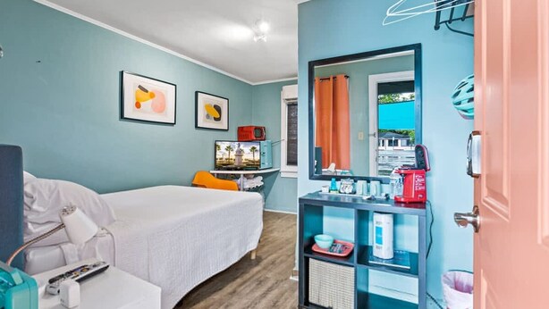 A hotel room with a bed, a desk with a computer, a chair, a TV, a mirror, and a shelf with various items. - Travel Back to the 50s- Renovated Motel Gem (St. Augustine)