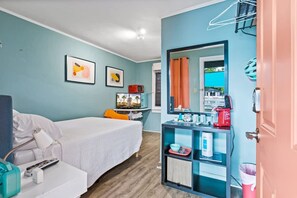 Habitación de hotel con cama, escritorio con computadora, silla, televisor, espejo y estante con varios objetos.