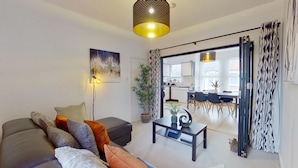 Ein modernes Wohnzimmer mit einer Couch, einem Couchtisch, einer Lampe und einem Essbereich, der durch eine offene Tür sichtbar ist.