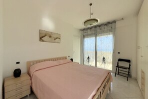 Ein Schlafzimmer mit Bett, Nachttisch, Sessel, einem Fenster mit Aussicht und einer Deckenleuchte.