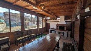 Un intérieur rustique en bois, avec une grande table et des bancs, une cheminée en pierre et de grandes fenêtres donnant sur l’extérieur.