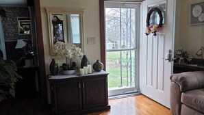 A living room with a wooden floor, a grey rug, a dark wooden cabinet, a white door with a wreath, and a window with blinds.