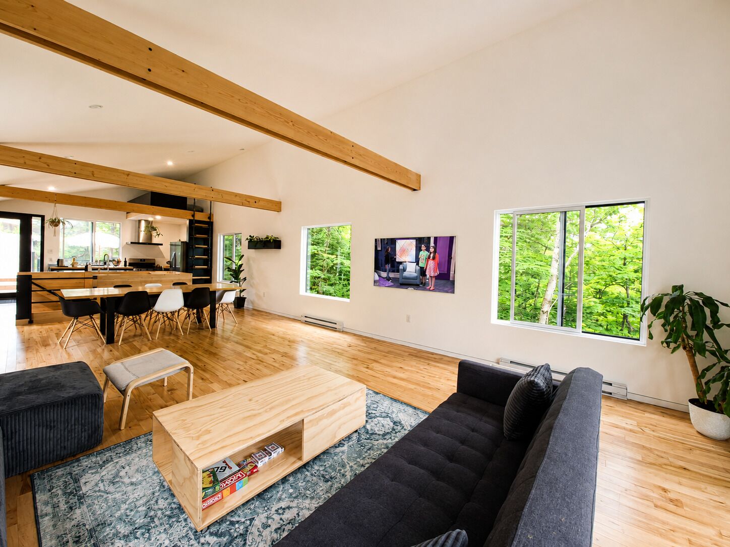 En rummelig opholdsstue med en mørk sofa, et træsofa bord og et tæppe. I baggrunden er der et køkken med træbjælker, og store vinduer med udsigt til grøn vegetation.