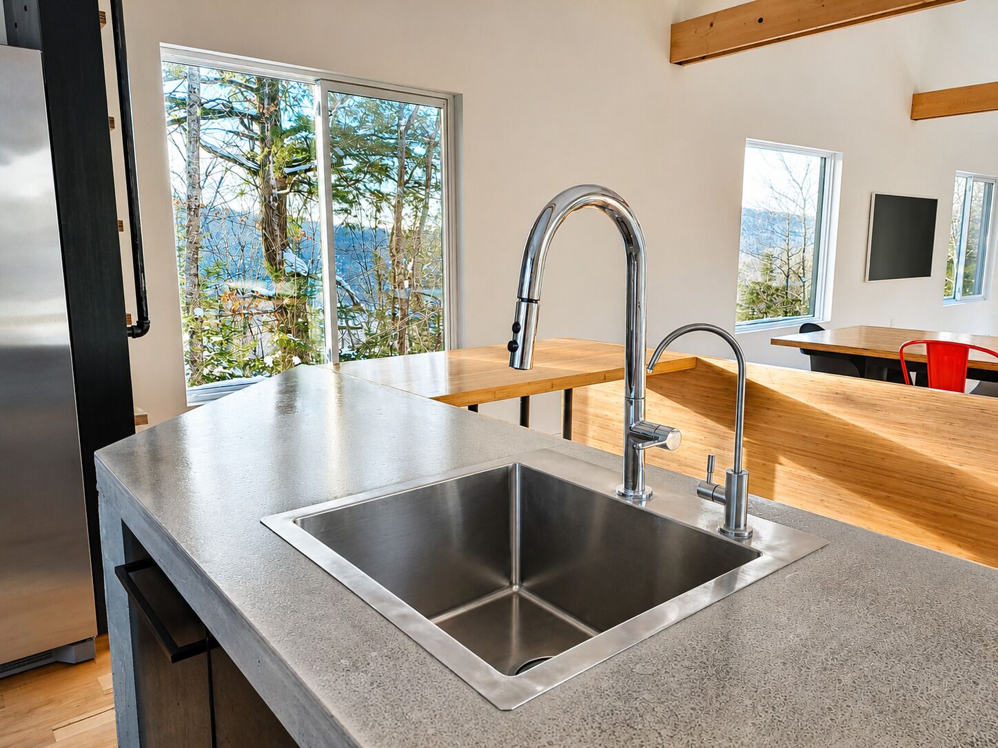 A modern kitchen with a stainless steel sink, wooden countertop, and a view of trees through large windows.