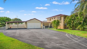 A two-story building with a covered parking area, a garage, and a lawn with palm trees. - Comfortable 2BR coastal unit with WiFi, BBQ area, parking & linen included, close to beach (Evans Head)