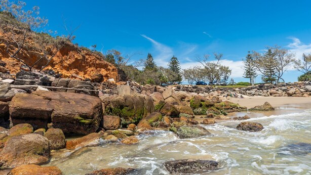 Una playa rocosa con aguas transparentes, árboles y una zona arenosa.