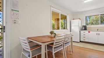 A kitchen with a wooden table and chairs, a refrigerator, and a window with a view of greenery.